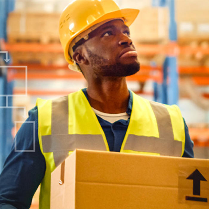 Man wearing hard helmet and high visibility vest holding a box standing in a warehouse