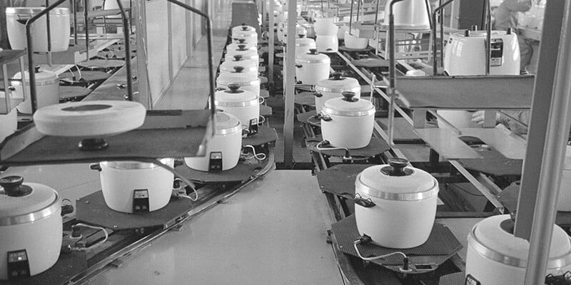 A historical photo of an assembly line of white rice cooker appliances.