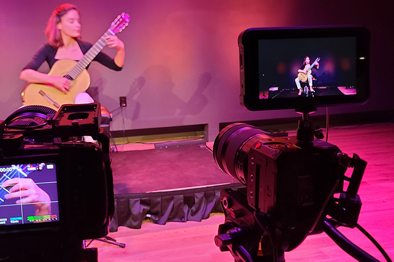 BS1H, on right side of frame, shoots a classical guitar performance with Blackmagic Pocket Cinema Camera on right side of frame.