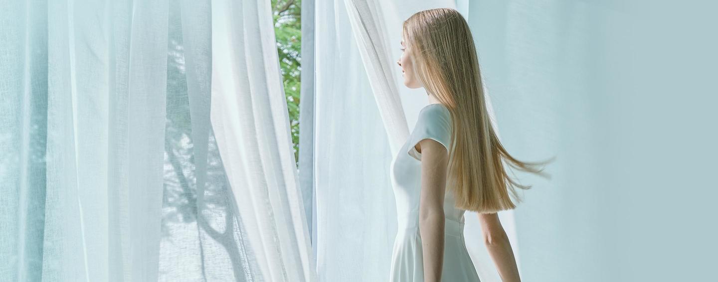 A woman, in white, stands by an open window and breathes in the fresh air as it blows through her white blinds.