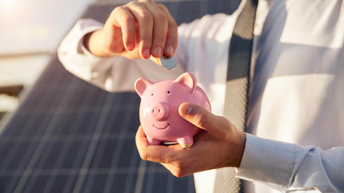 Man putting money into piggy bank near solar panel