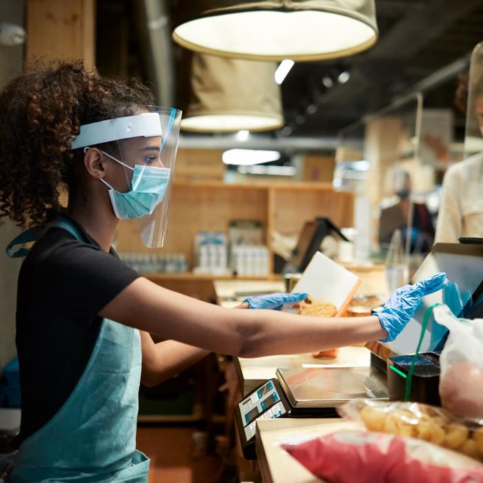 a cashier in protective gear rings up a customer at a grocery store