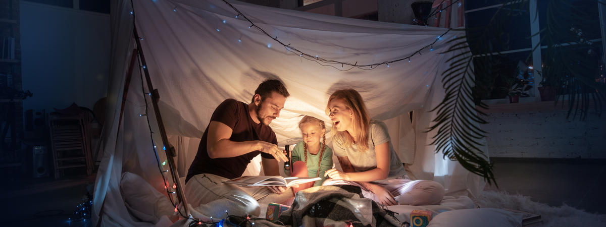 Caucasian family sitting in a teepee, reading stories with the flashlight in dark room with toys and pillows. Caucasian models. Home comfort, family, love, Christmas holidays, storytelling time.